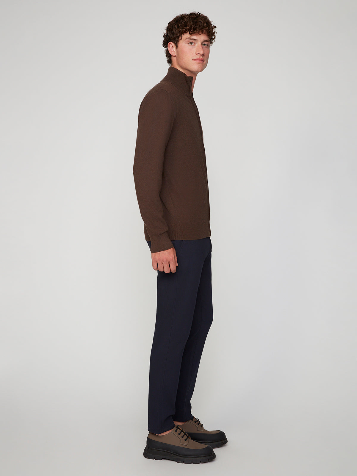 A man stands in profile against a plain light gray background, wearing a brown zip-up sweater, PROJEK RAW MEN’S RELAXED BUNGEE TWILL CHINO PANT, and brown lace-up shoes.