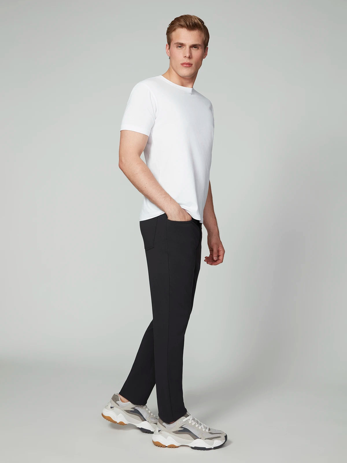 A young man stands in a studio wearing PROJEK RAW MENS 5-POCKET BUNGEE TWILL PANT in black, paired with a plain white t-shirt and gray chunky sneakers. He has short light brown hair and poses with one hand in his pocket, looking to the side.