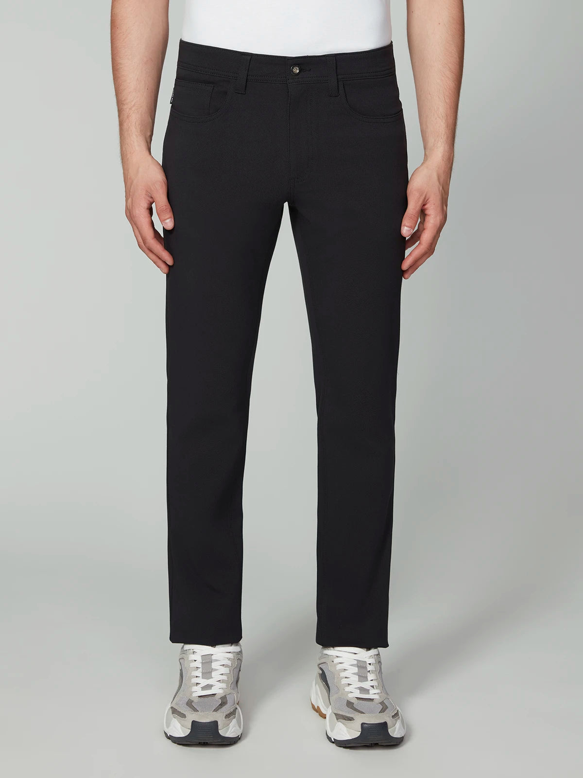 A person wearing PROJEK RAW MENS 5-POCKET BUNGEE TWILL PANT in black, a white t-shirt, and gray sneakers stands facing forward against a plain light gray background.