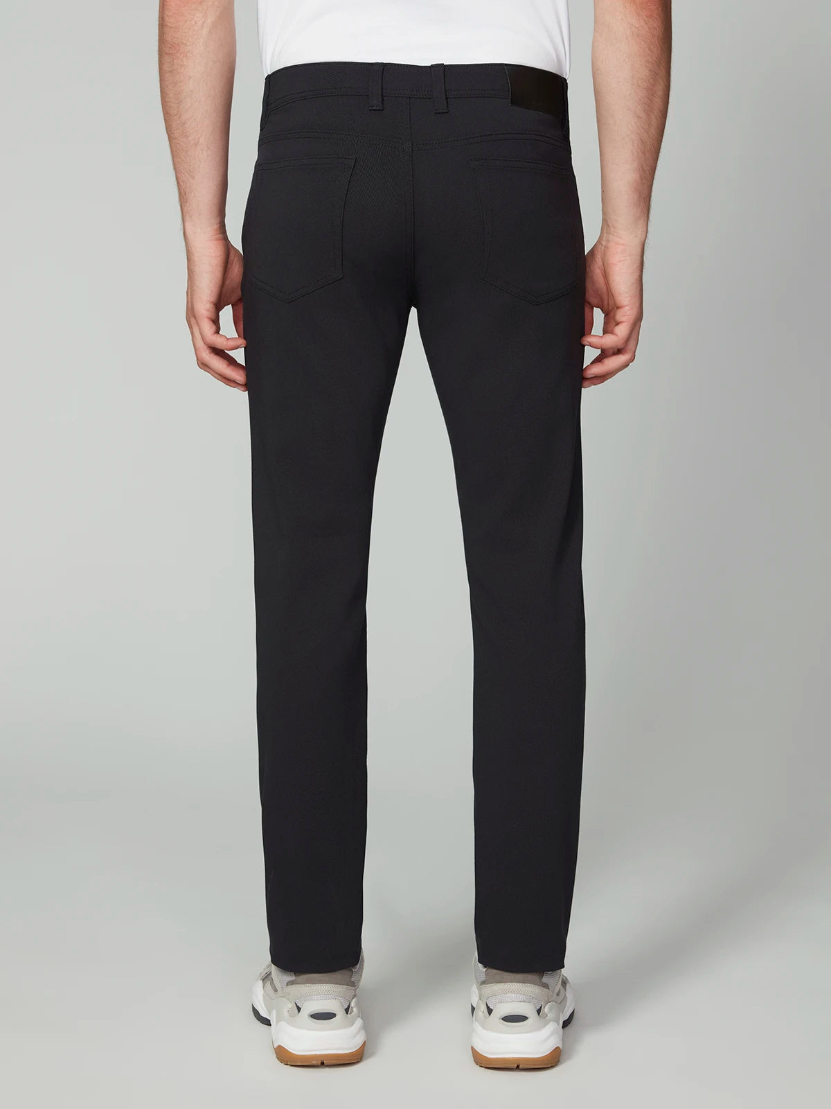 A person faces away from the camera, wearing PROJEK RAW MENS 5-POCKET BUNGEE TWILL PANT in black, paired with white sneakers and a white shirt against a plain light gray background.