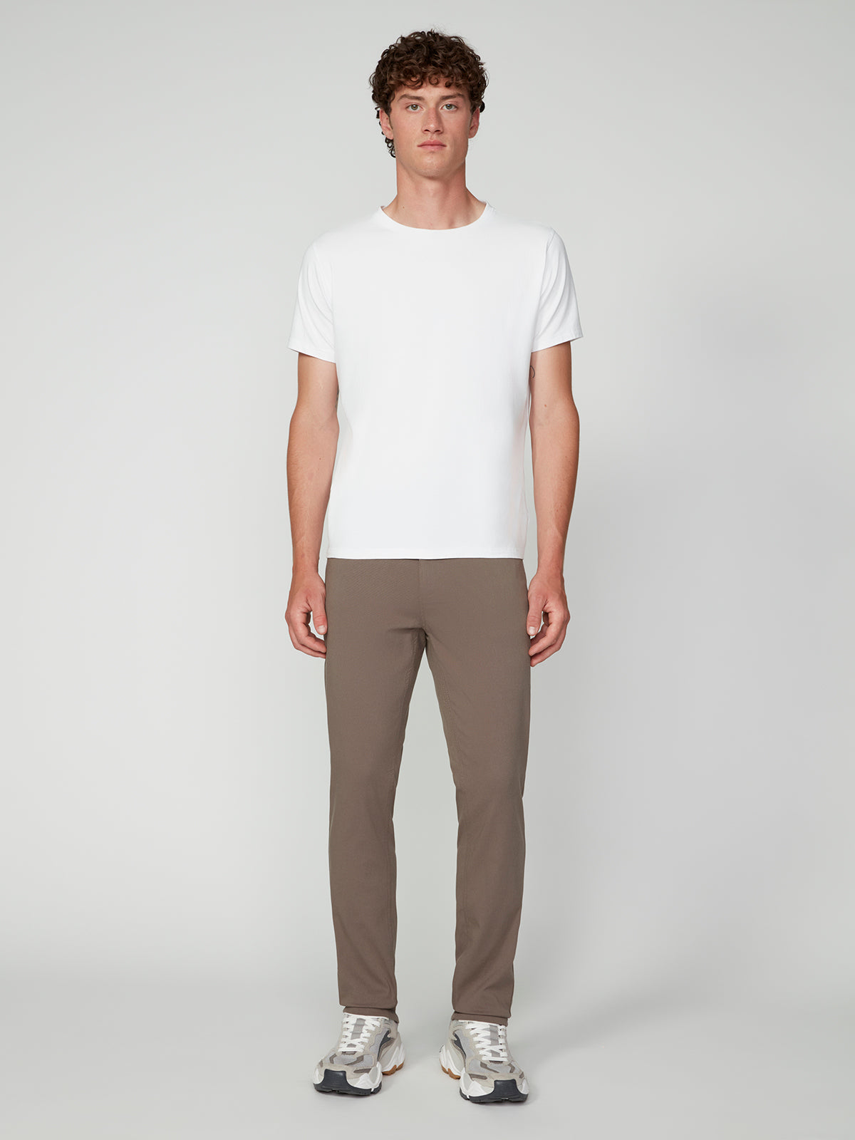 A man with curly hair faces forward in a plain white t-shirt, PROJEK RAW MENS 5-POCKET BUNGEE TWILL PANT in taupe, and white sneakers against a light gray background.