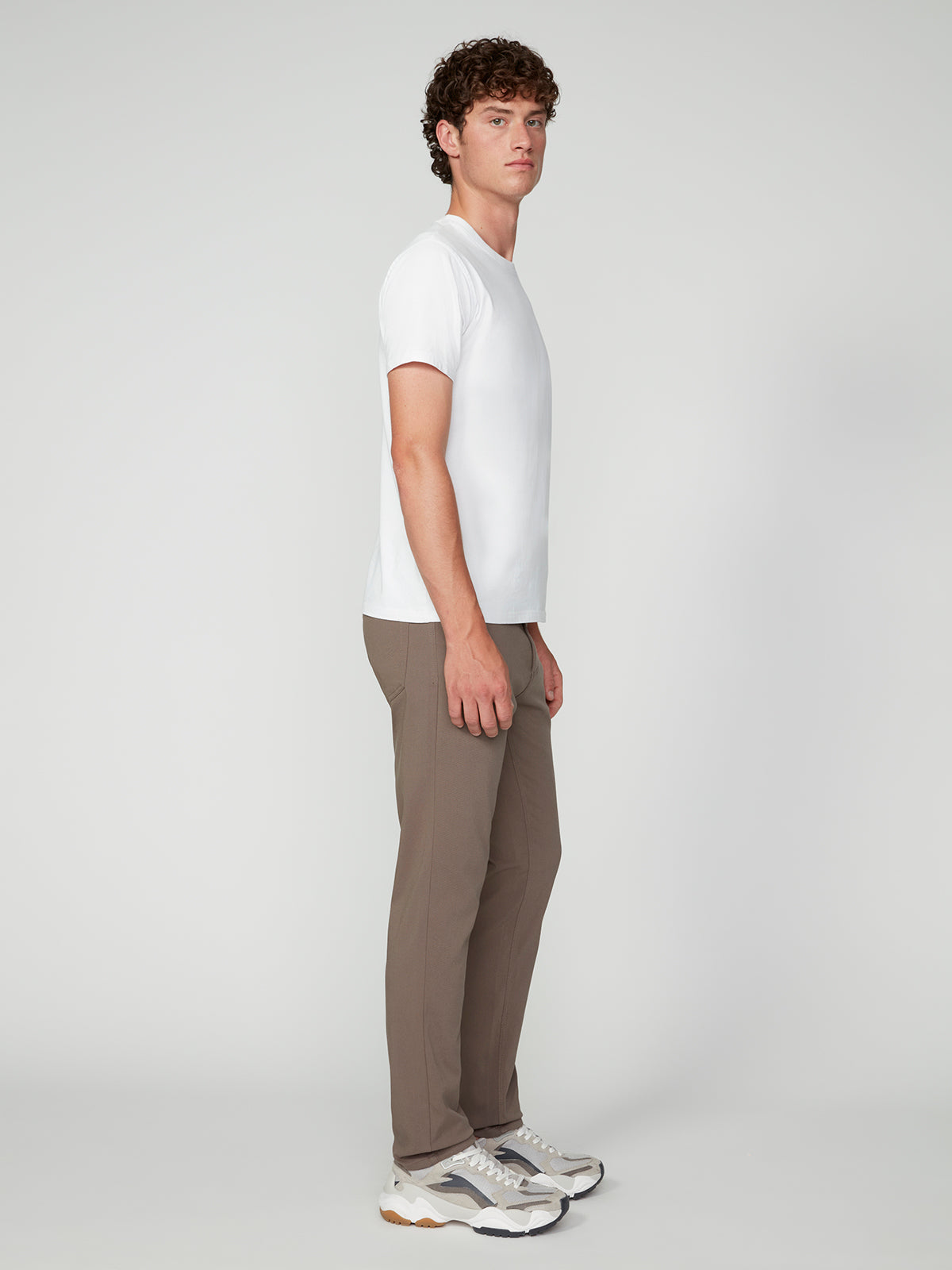 A young man with curly hair stands sideways in a plain white t-shirt, white sneakers, and PROJEK RAW MENS 5-POCKET BUNGEE TWILL PANT. The background is plain and light gray.