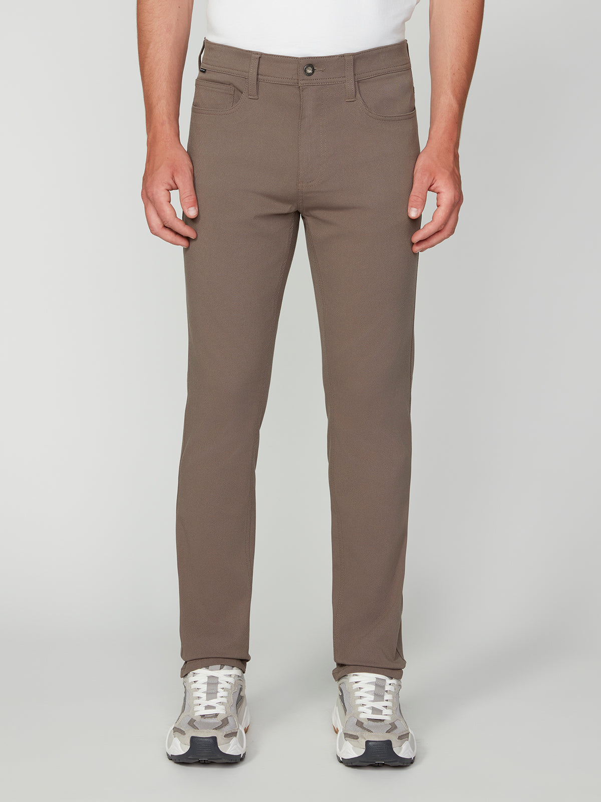 A person in PROJEK RAW MENS 5-POCKET BUNGEE TWILL PANT (taupe), a white shirt, and light gray sneakers stands facing forward against a plain light background.
