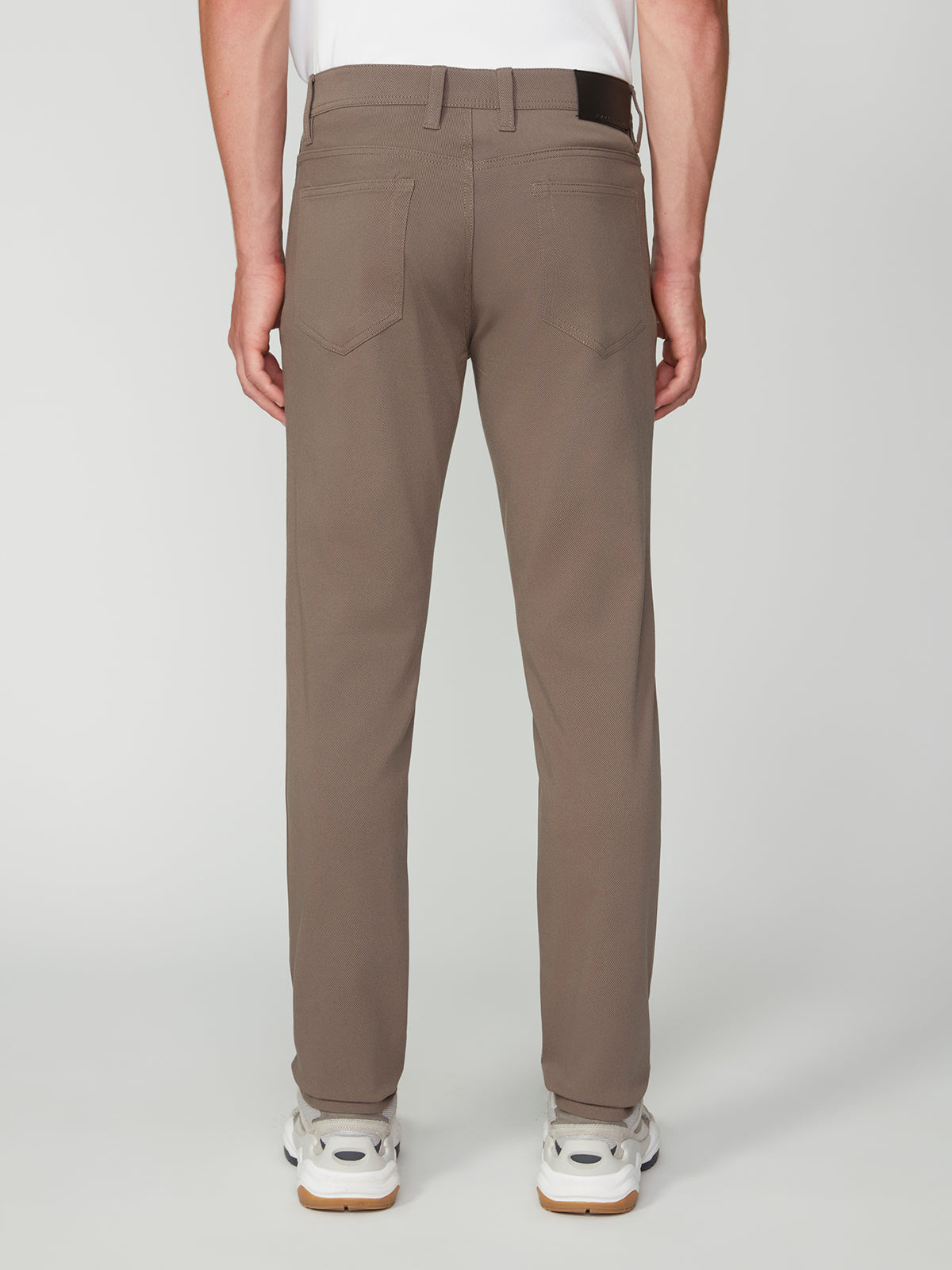 A person stands facing away from the camera, wearing PROJEK RAW MENS 5-POCKET BUNGEE TWILL PANT in taupe, paired with a white shirt and white sneakers against a plain light background.