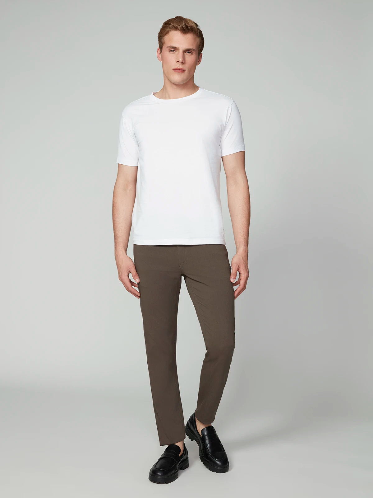 A man faces forward with a neutral expression, wearing a white short-sleeve t-shirt, black shoes, and PROJEK RAW MENS 5-POCKET BUNGEE TWILL PANT in brown. His hands rest relaxed by his sides against a plain background.
