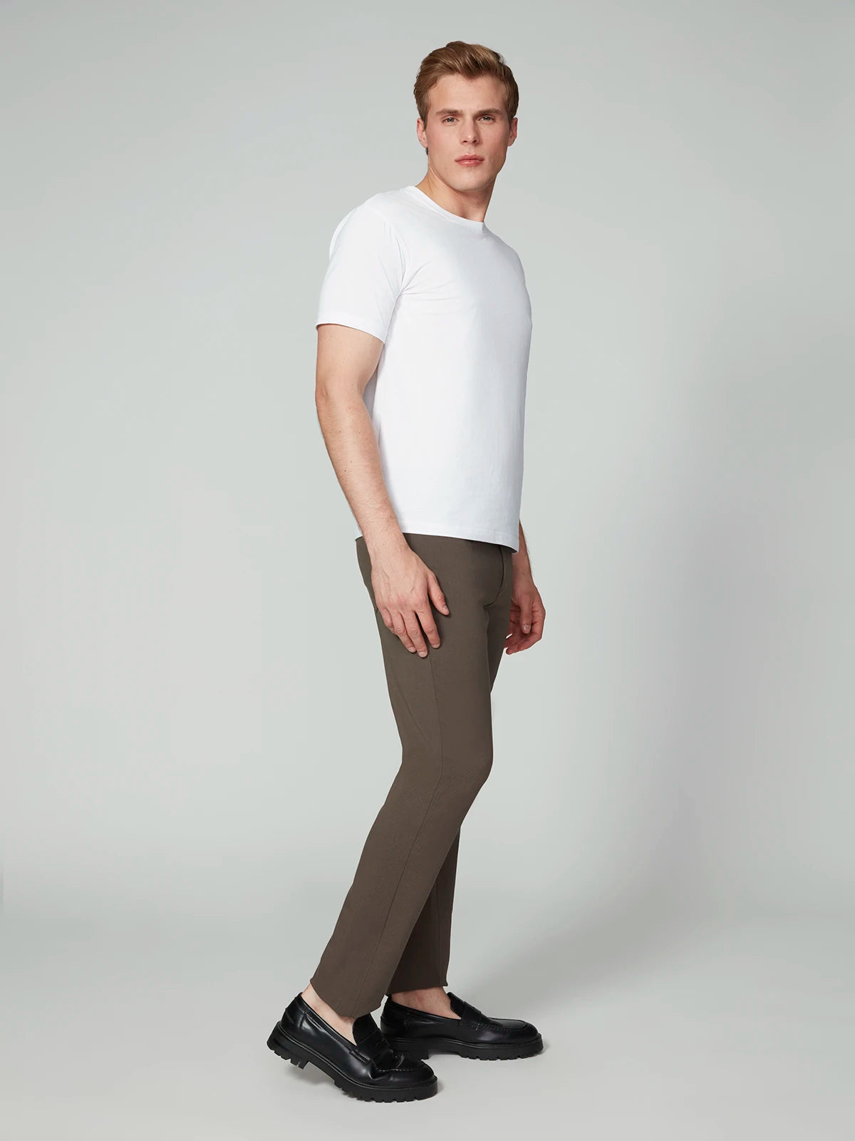 A man stands in a studio, facing sideways with his head turned to the camera, wearing a plain white t-shirt, PROJEK RAW MENS 5-POCKET BUNGEE TWILL PANT, and black loafers against a light grey background.