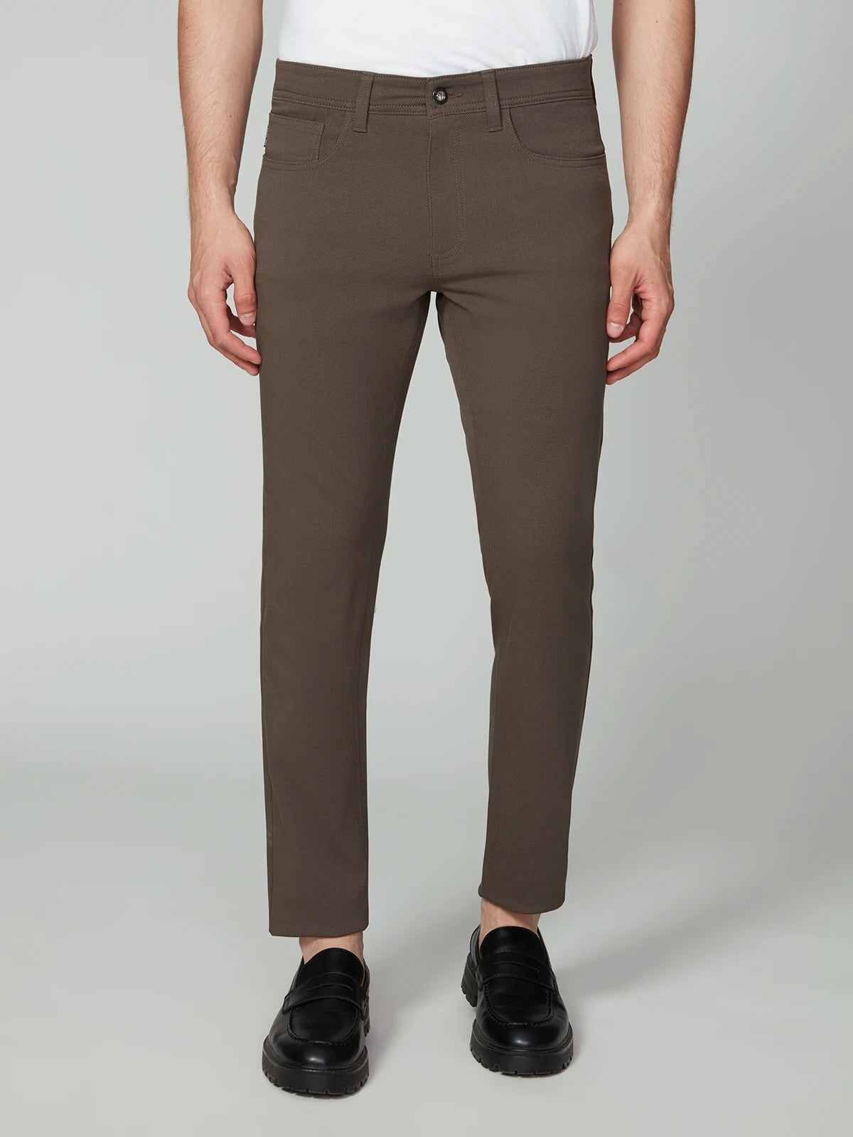 A person stands facing forward in PROJEK RAW MENS 5-POCKET BUNGEE TWILL PANT (brown), a white shirt, and black loafers. The image is cropped at the shoulders, focusing on the pants and shoes against a plain light backdrop.