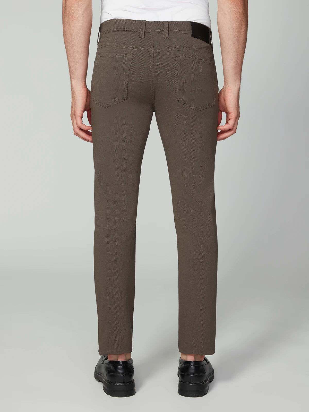 A person wearing PROJEK RAW MENS 5-POCKET BUNGEE TWILL PANT in brown, paired with a white shirt and black shoes, stands facing away from the camera, showing the back view of the outfit against a plain light background.