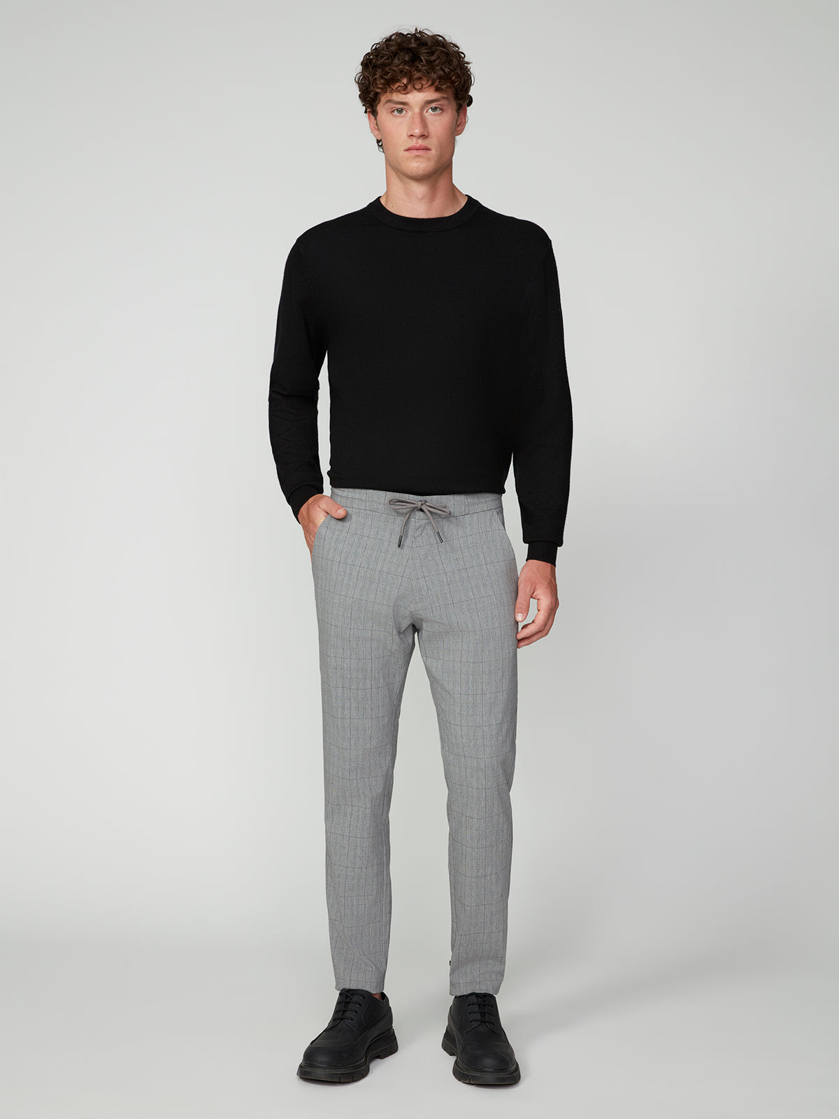 A man faces forward wearing PROJEK RAW MEN’S PLAID BUNGEE DRAWSTRING PANT, paired with a black long-sleeve shirt and black dress shoes, against a plain light gray background.