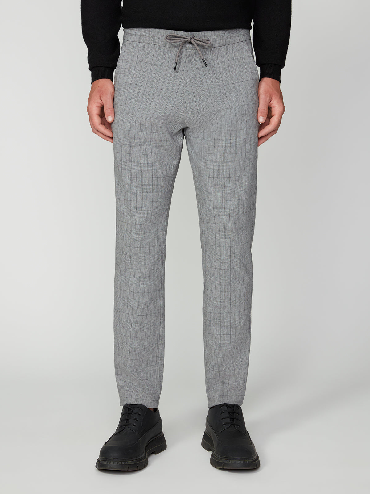 Wearing PROJEK RAW MEN’S PLAID BUNGEE DRAWSTRING PANT in light gray with a black sweater and lace-up shoes, a person stands against a plain light background.