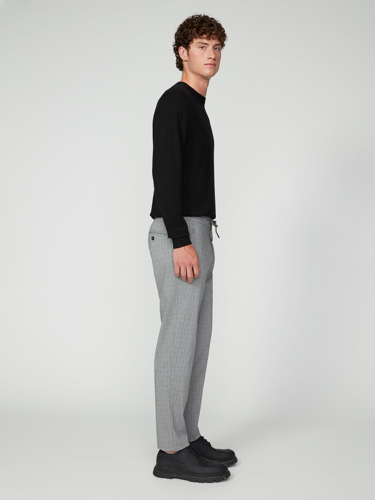Man standing sideways against a plain white background, wearing a black long-sleeve shirt, PROJEK RAW MEN’S PLAID BUNGEE DRAWSTRING PANT, and black shoes, looking ahead with a neutral expression.