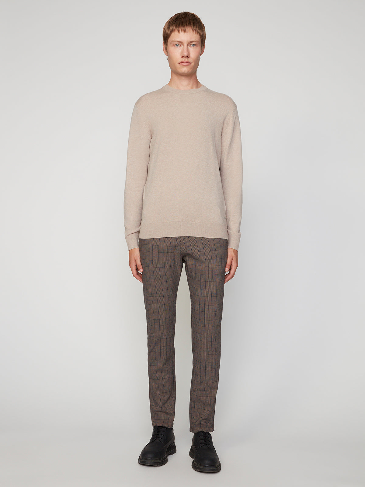 A person stands facing forward against a plain light background, wearing a beige long-sleeve sweater, PROJEK RAW MENS SOLID BUNGEE RELAXED 5-POCKET PANT, and black shoes.