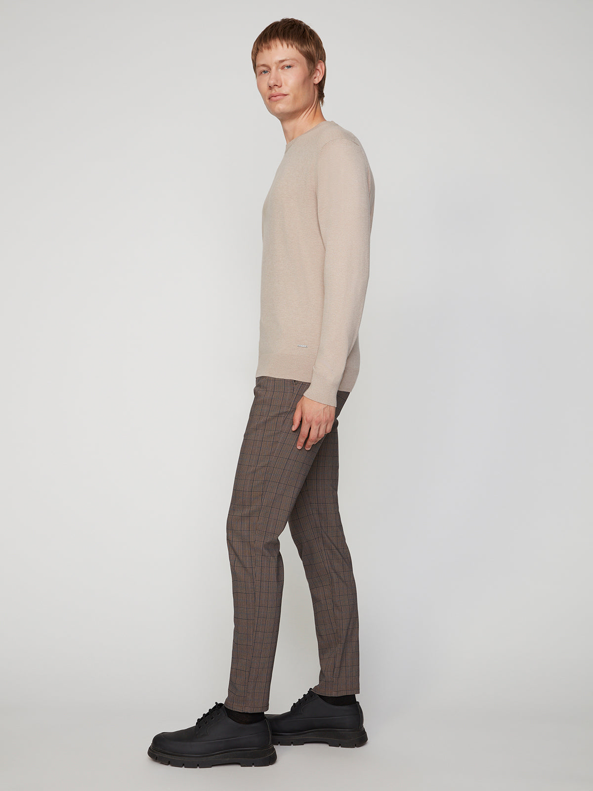 A person with short light brown hair stands sideways in a beige long-sleeve sweater, PROJEK RAW MENS SOLID BUNGEE RELAXED 5-POCKET PANT, and black chunky shoes against a plain light gray background, highlighting modern style.