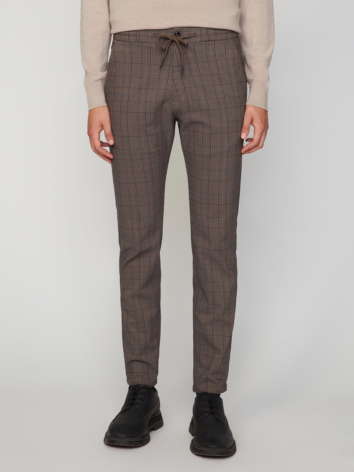 A man wearing PROJEK RAW MENS SOLID BUNGEE RELAXED 5-POCKET PANT stands against a light background in a beige sweater and black lace-up shoes, highlighting the relaxed fit and modern style of these pants.