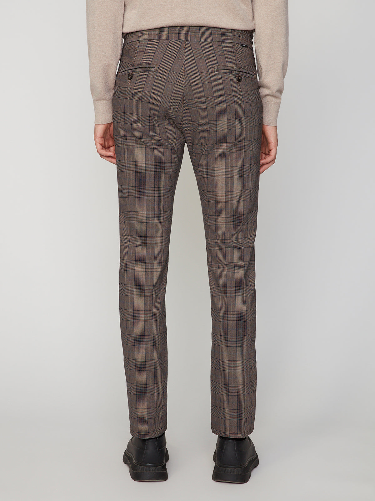 A person faces away wearing PROJEK RAW MENS SOLID BUNGEE RELAXED 5-POCKET PANT in brown with buttoned back pockets, a beige sweater, and black shoes. The pants have a tailored fit and the background is plain and light-colored.