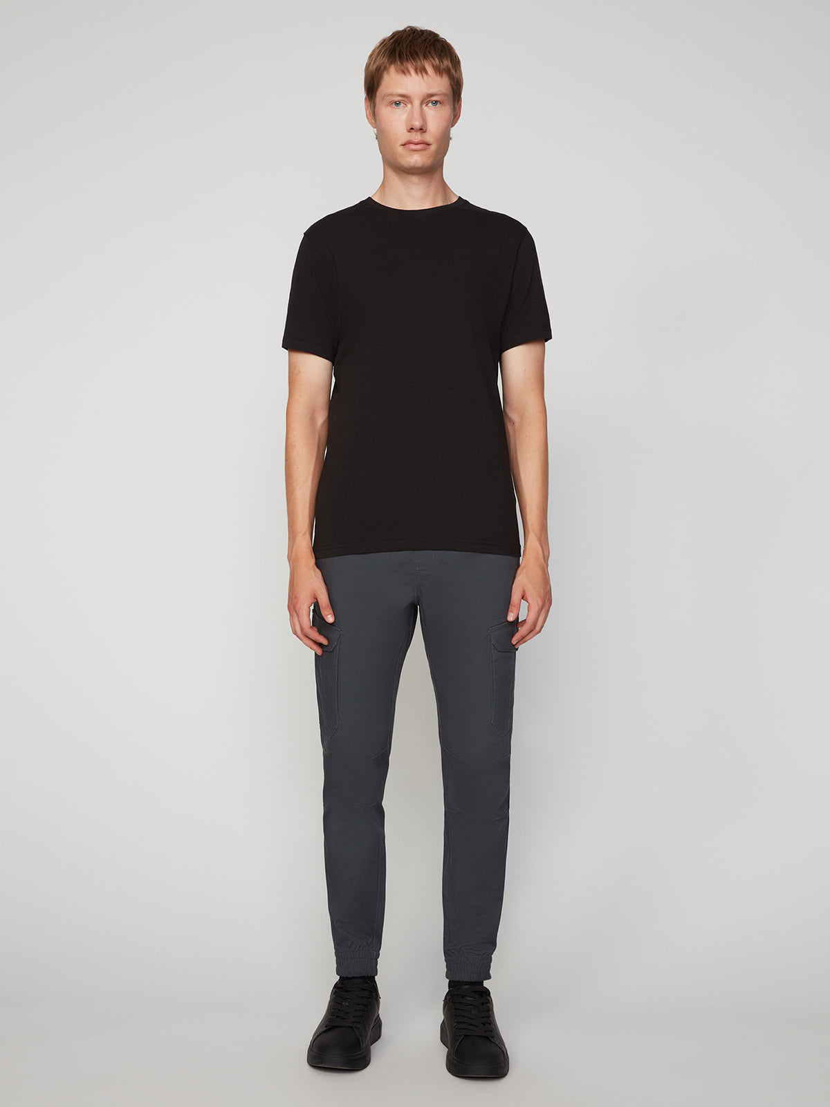 A person stands against a plain light background, wearing a black short-sleeve t-shirt, PROJEK RAW MEN’S CANVAS BUNGEE PULL-ON CARGO JOGGER in dark gray, and black shoes.