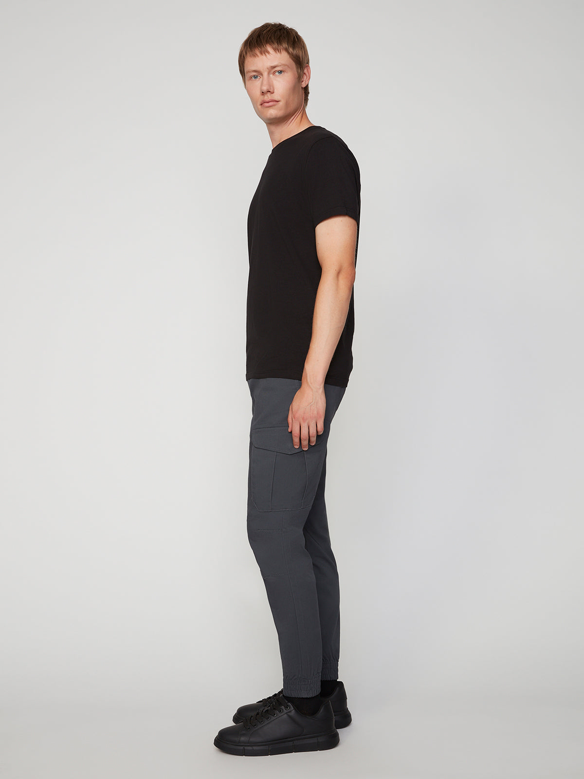 A man stands sideways against a light background, wearing a black t-shirt, PROJEK RAW MEN’S CANVAS BUNGEE PULL-ON CARGO JOGGER with pockets, and black sneakers. He faces the camera slightly with a neutral expression.