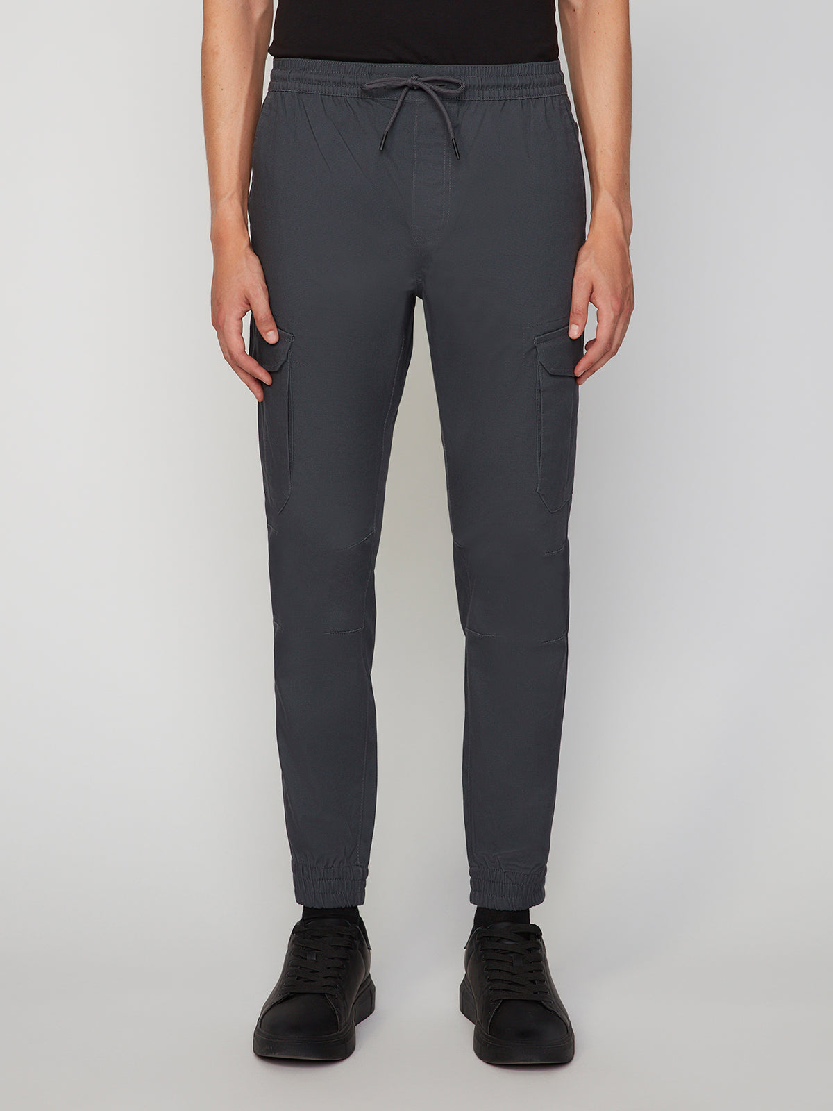 A person wears PROJEK RAW MEN’S CANVAS BUNGEE PULL-ON CARGO JOGGER in dark gray, featuring an elastic waistband, drawstring, side pockets, and paired with black shoes, standing before a plain light background.
