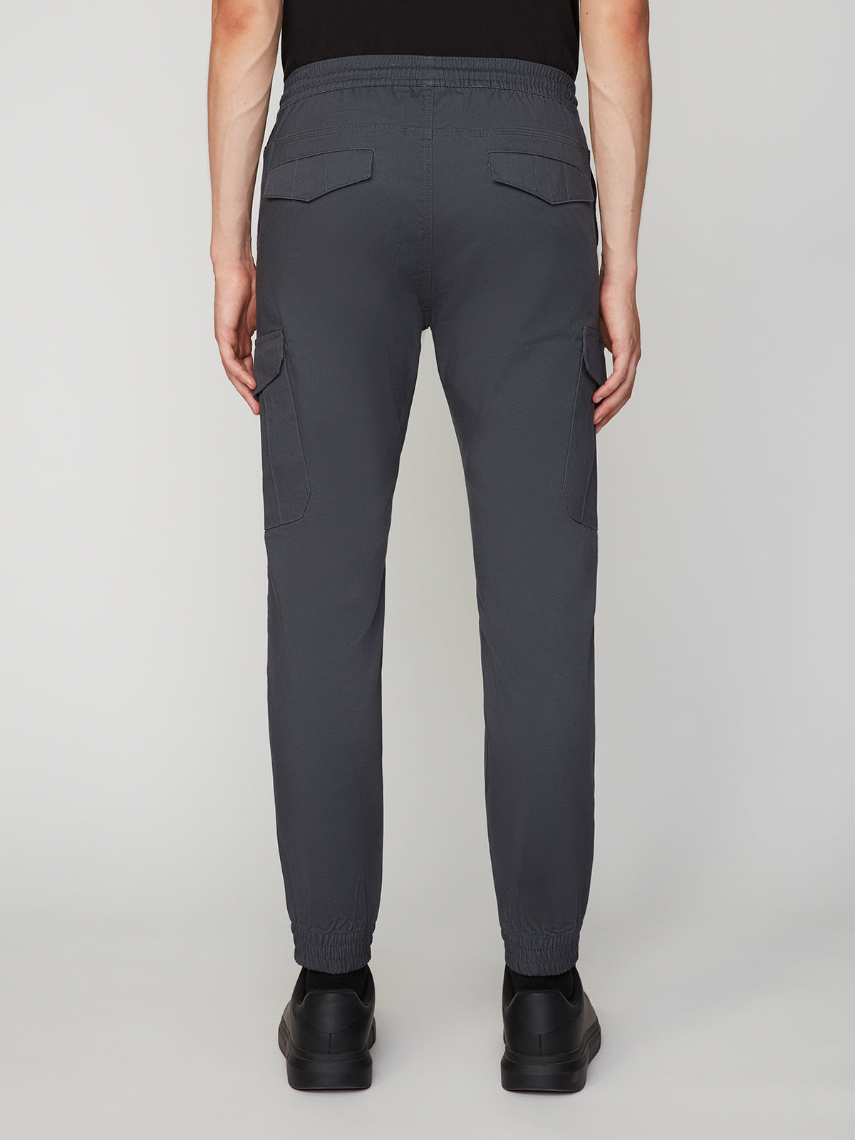 A person wearing PROJEK RAW MEN’S CANVAS BUNGEE PULL-ON CARGO JOGGER in dark gray and black shoes is shown from the back against a plain light background. The joggers have flap back pockets and large side cargo pockets.