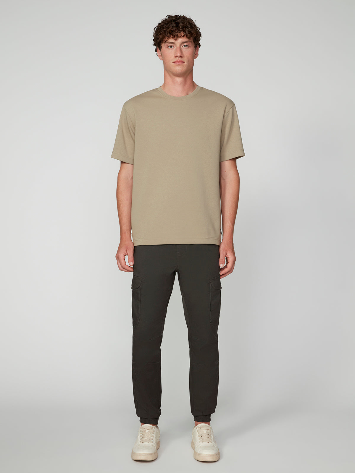 A young man with curly brown hair stands against a gray background, wearing a beige short-sleeve T-shirt, PROJEK RAW MEN’S CANVAS BUNGEE PULL-ON CARGO JOGGER, and white sneakers. He faces the camera with a neutral expression.