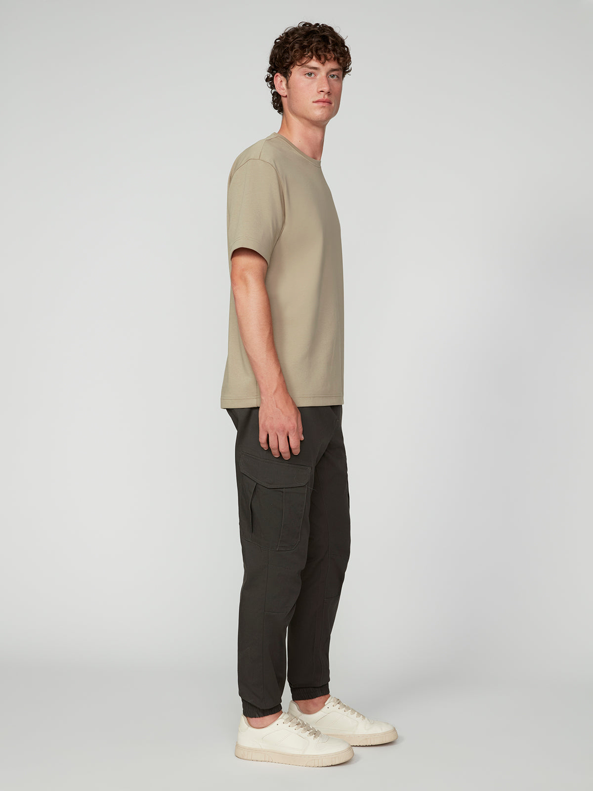 A young man with curly hair stands sideways, wearing a beige t-shirt, PROJEK RAW MEN’S CANVAS BUNGEE PULL-ON CARGO JOGGER, and white sneakers against a plain light gray background.