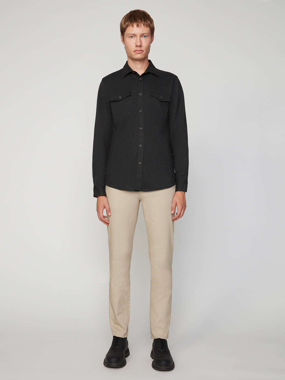 A man stands facing forward against a plain background, wearing a black button-up shirt, PROJEK RAW MENS CANVAS BUNGEE 5-POCKET PANT in beige, and black shoes.