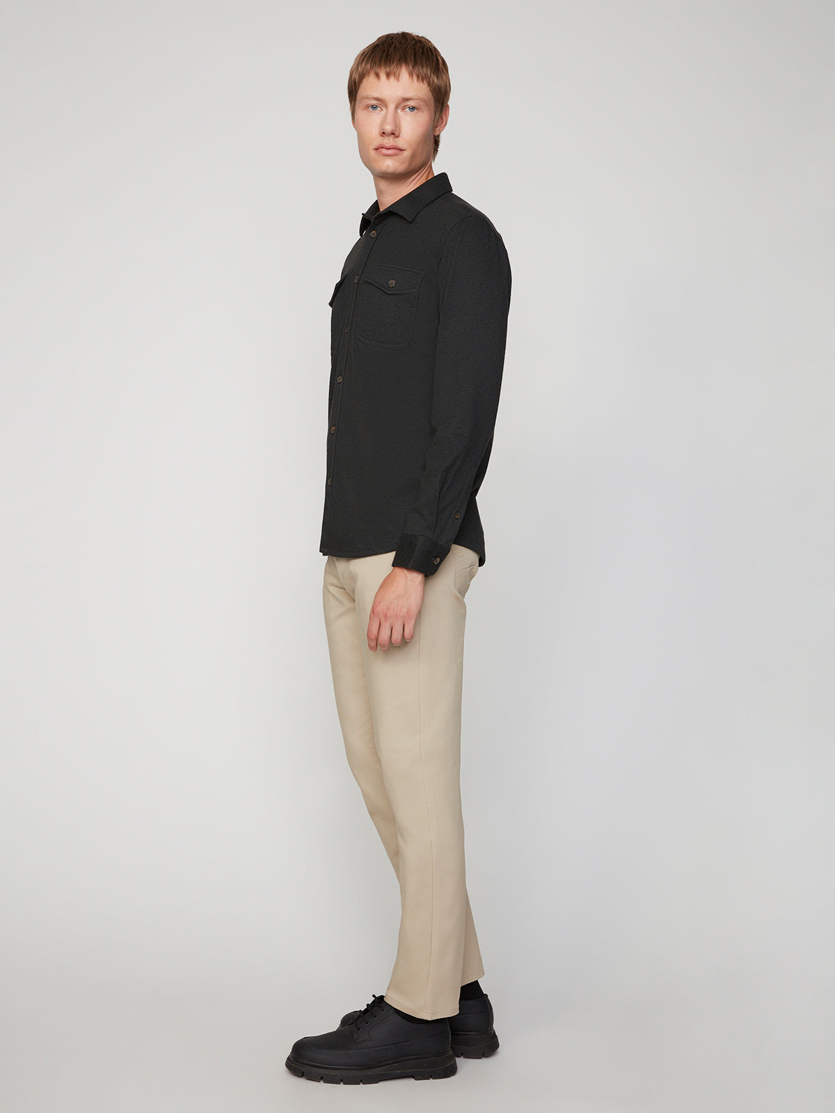 A man stands sideways against a plain light background, wearing a black button-up shirt, PROJEK RAW MENS CANVAS BUNGEE 5-POCKET PANT in beige, and black shoes. He looks at the camera with a neutral expression.