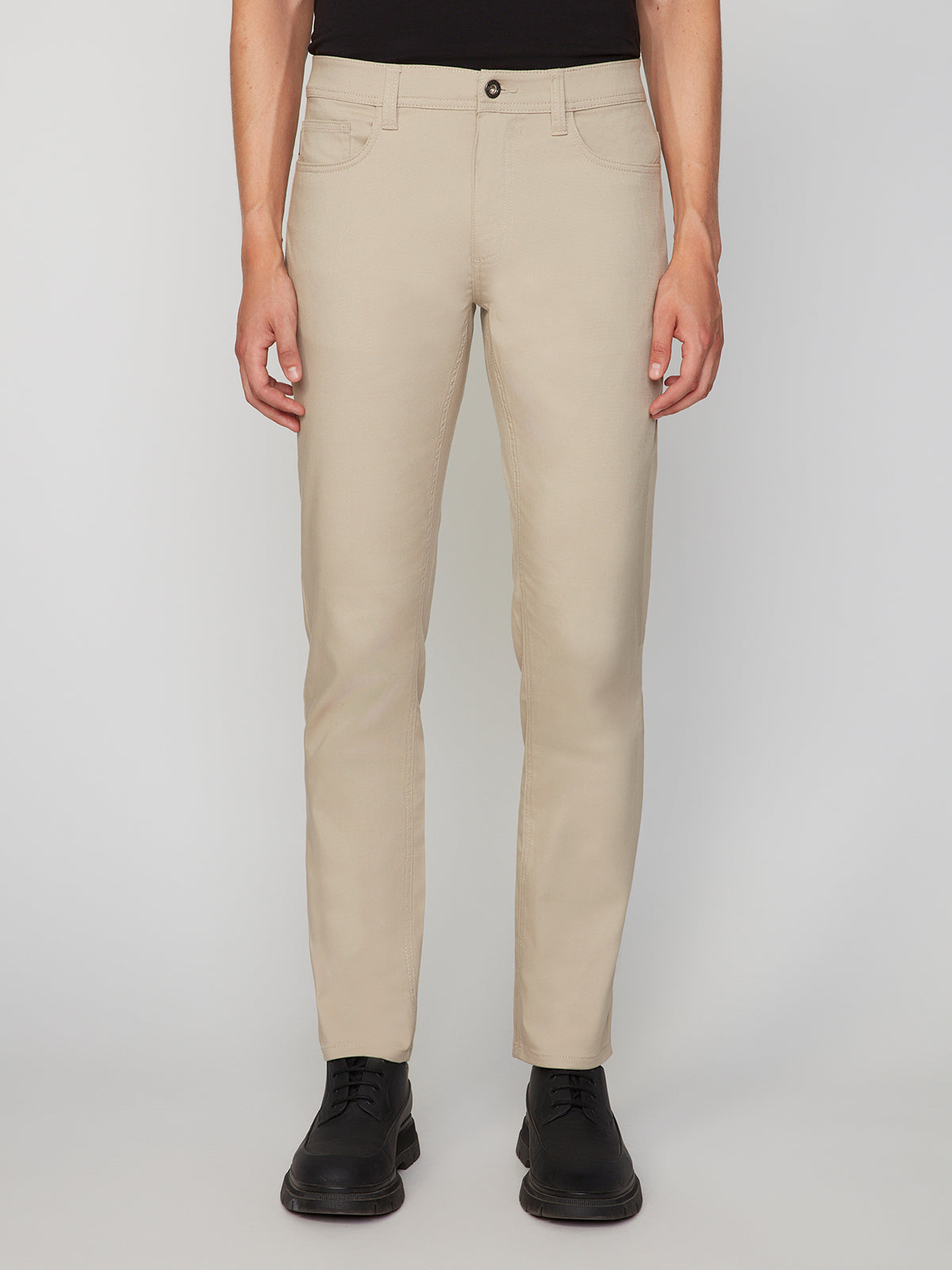 A person wears PROJEK RAW MENS CANVAS BUNGEE 5-POCKET PANT in beige, paired with a black top and black shoes, standing against a plain light gray background.