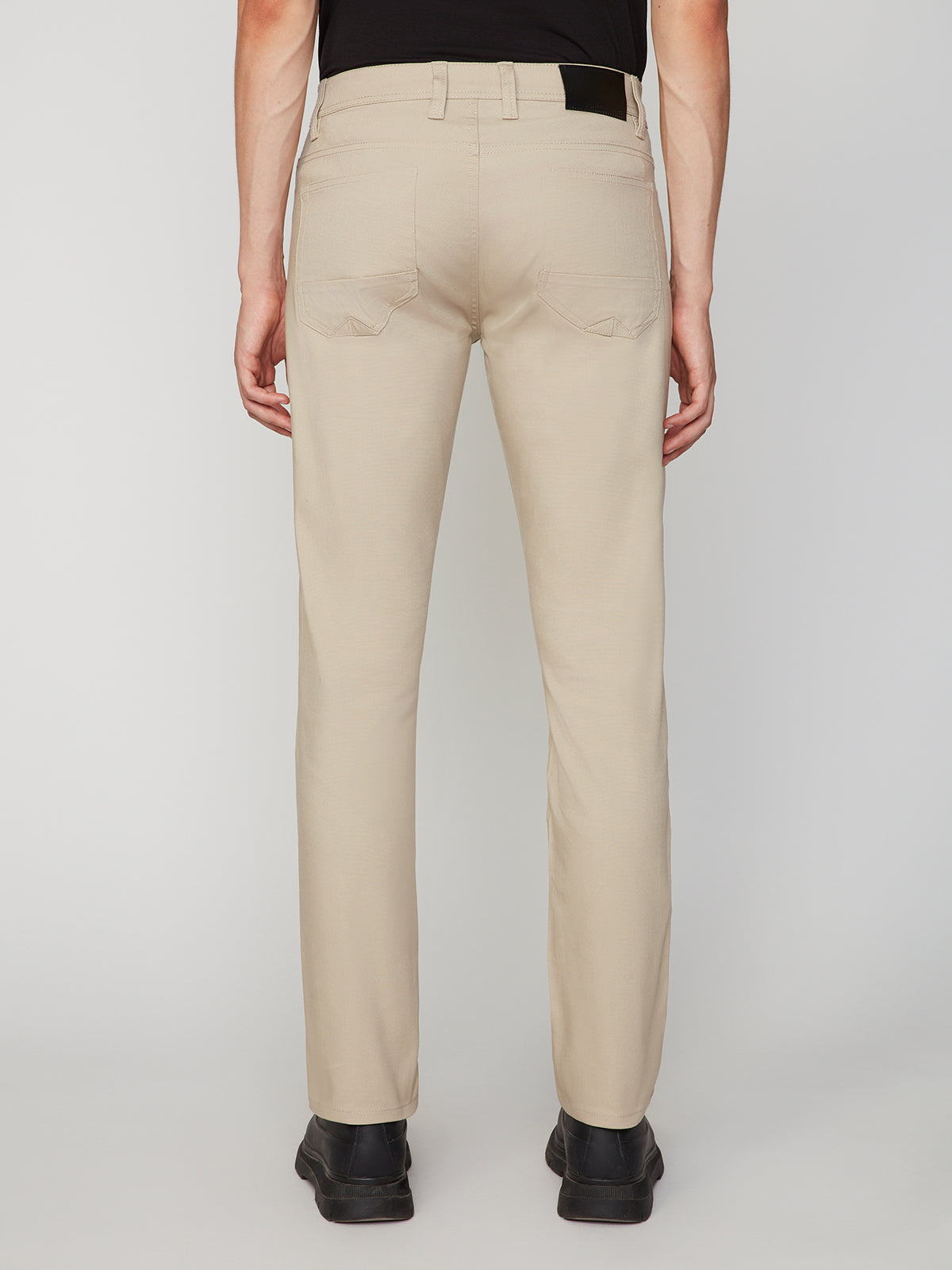 A person stands facing away, wearing PROJEK RAW MENS CANVAS BUNGEE 5-POCKET PANT in beige with straight legs and stitched back pockets. Paired with a black shirt and shoes, these pants stand out against a plain, light background.
