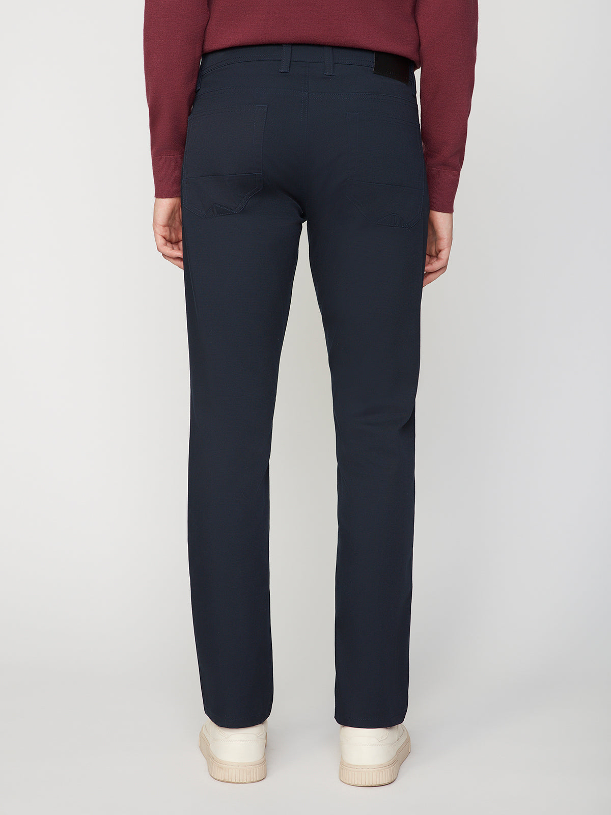 A person stands facing away from the camera, wearing a maroon sweater, PROJEK RAW MENS CANVAS BUNGEE 5-POCKET PANT in navy blue, and white sneakers against a plain light background.