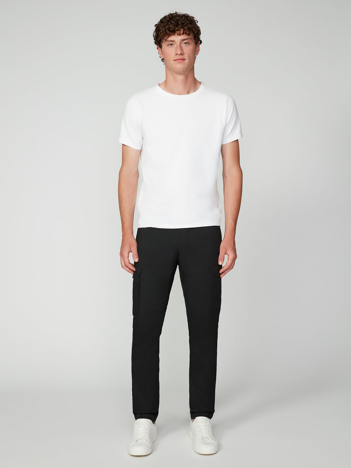 A young man with curly brown hair wears a white tee, PROJEK RAW MENS CANVAS BUNGEE RELAXED CARGO PANT with side pockets, and white sneakers. He stands against a plain background facing forward with a neutral expression.