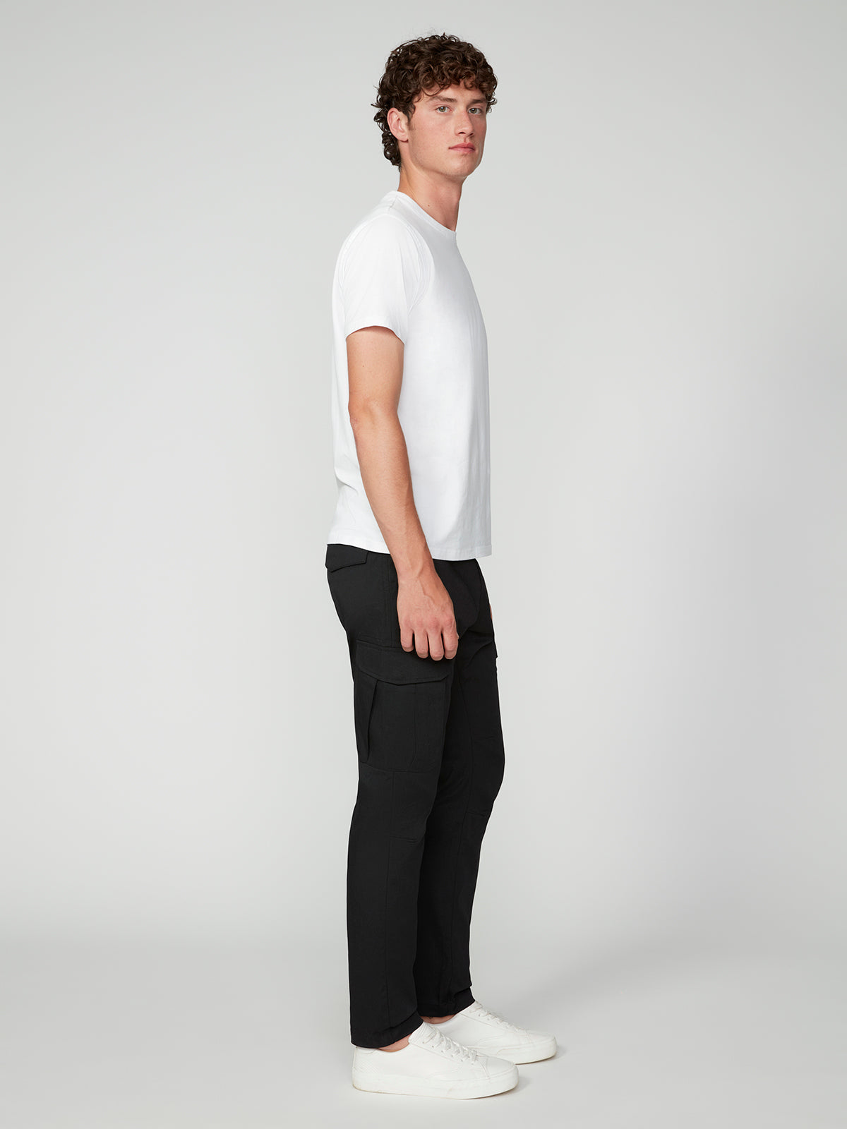 A young man with curly hair stands sideways against a light gray background, wearing a plain white T-shirt, PROJEK RAW MENS CANVAS BUNGEE RELAXED CARGO PANT, and white sneakers.