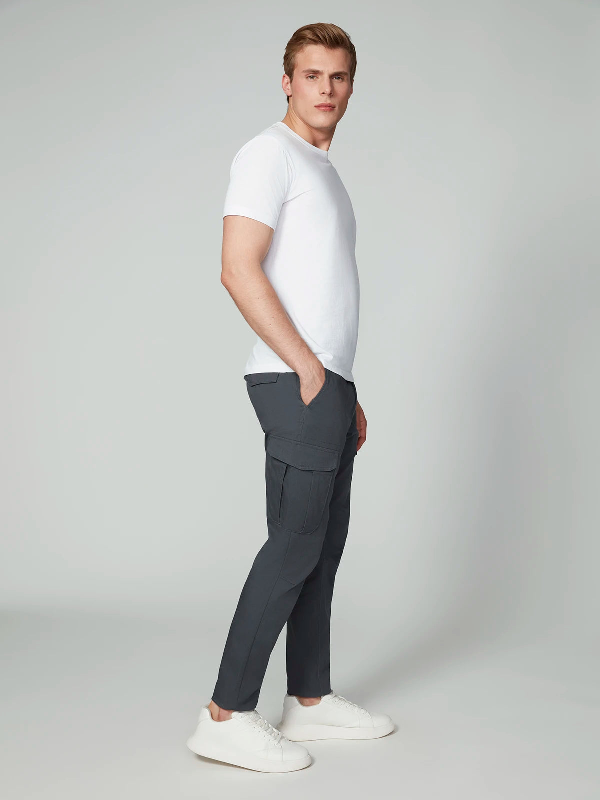 A man stands in a relaxed pose wearing a white t-shirt, white sneakers, and PROJEK RAW MENS CANVAS BUNGEE RELAXED CARGO PANT with pockets, against a light gray background.
