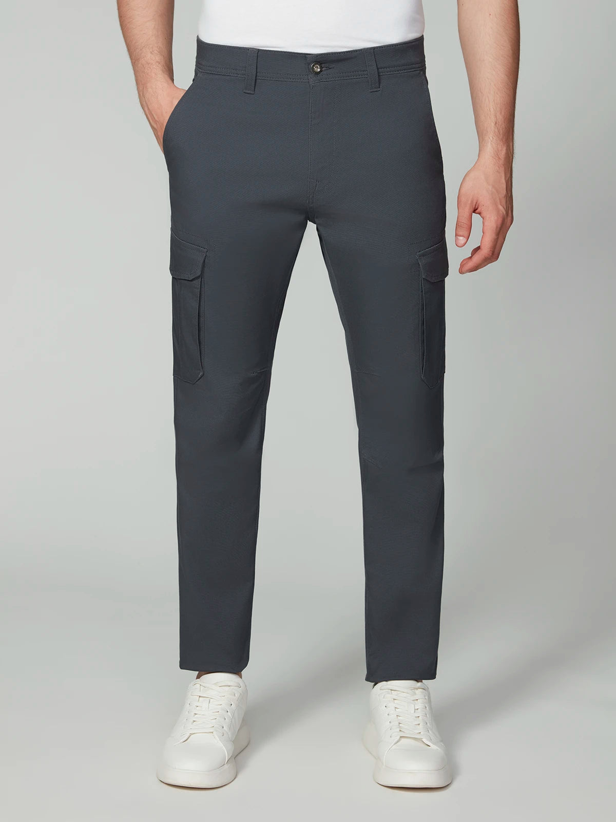 A person wearing PROJEK RAW MENS CANVAS BUNGEE RELAXED CARGO PANT in dark gray, paired with a white shirt and white sneakers, stands hands in pockets against a light gray background.