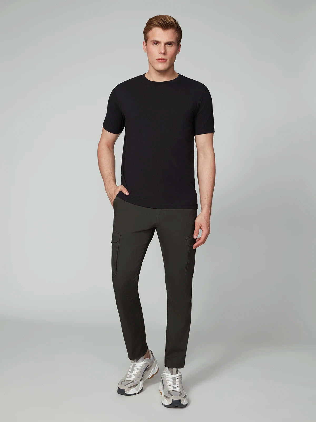 A man stands against a light gray background, wearing PROJEK RAW MENS CANVAS BUNGEE RELAXED CARGO PANT in dark gray, paired with a black tee and white-gray sneakers. One hand is in his pocket as he faces forward with a neutral expression.