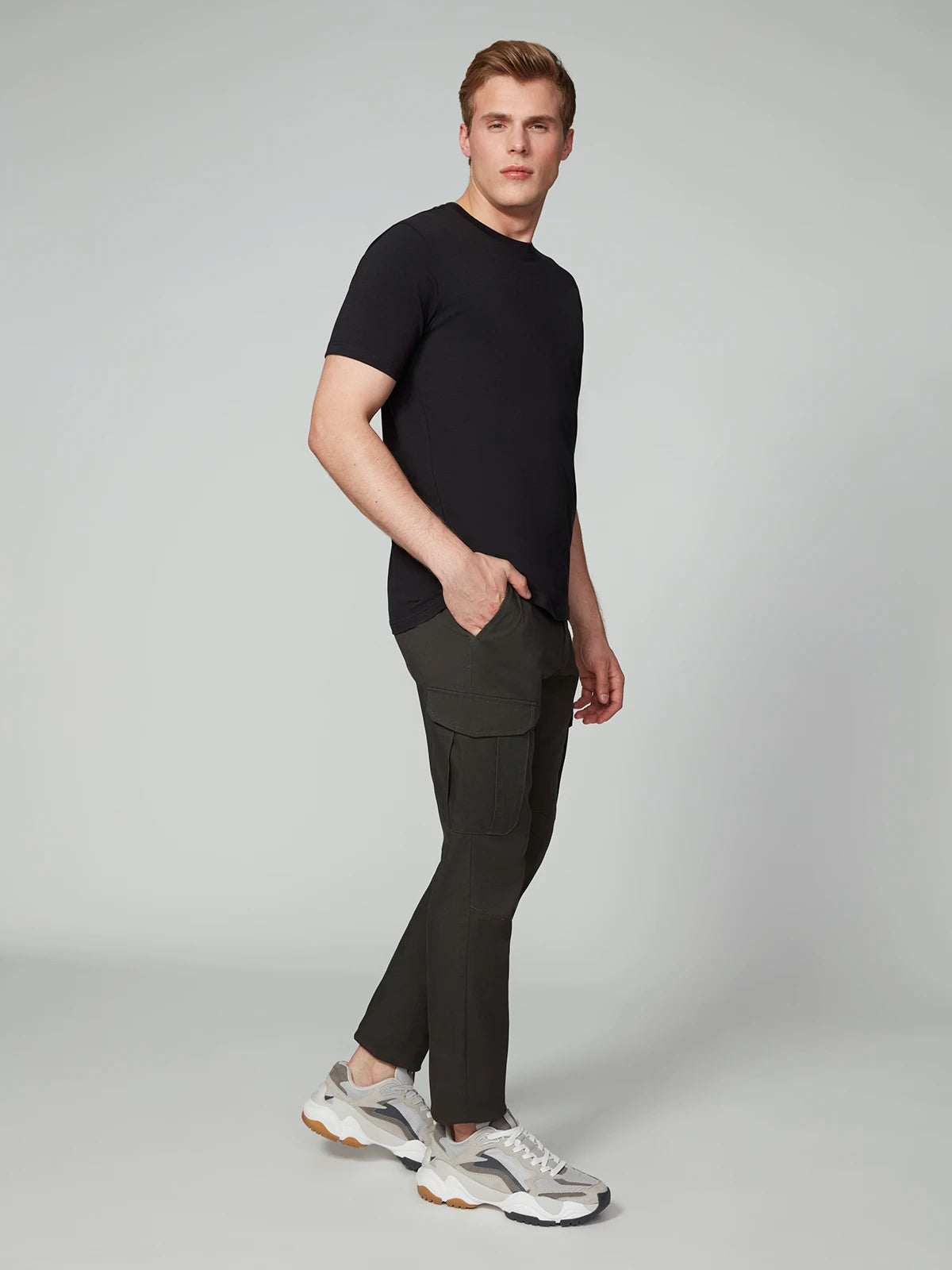 A person stands against a plain background, wearing PROJEK RAW MENS CANVAS BUNGEE RELAXED CARGO PANT in dark stretch canvas with a bungee waistband, styled with a black T-shirt and white sneakers with beige and gray accents. One hand is in their pocket.