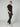 A person stands against a plain background, wearing PROJEK RAW MENS CANVAS BUNGEE RELAXED CARGO PANT in dark stretch canvas with a bungee waistband, styled with a black T-shirt and white sneakers with beige and gray accents. One hand is in their pocket.