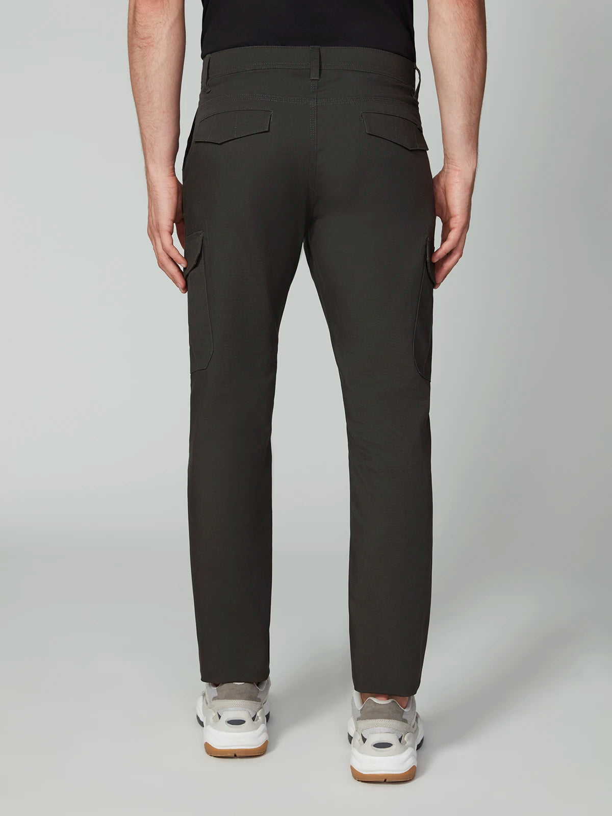 A person stands with their back to the camera in PROJEK RAW MENS CANVAS BUNGEE RELAXED CARGO PANT, featuring dark grey stretch canvas, flap back pockets, black side panels, bungee waistband, and white sneakers against a plain light background.
