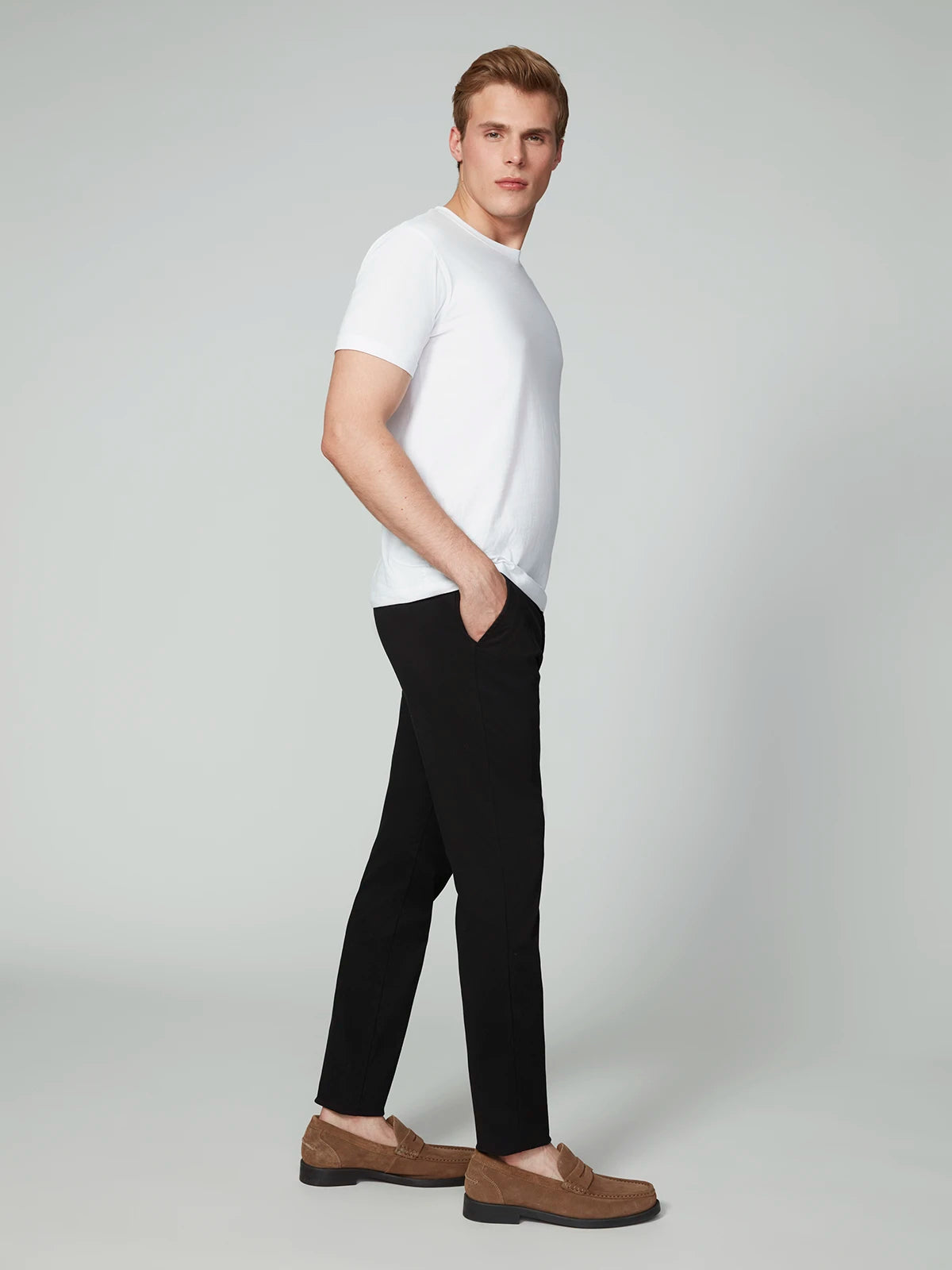 A man stands sideways with one hand in his pocket, wearing a white t-shirt, PROJEK RAW MENS CHINO SIGNATURE WOVEN PANT in a tailored fit, and brown loafers against a plain light background.