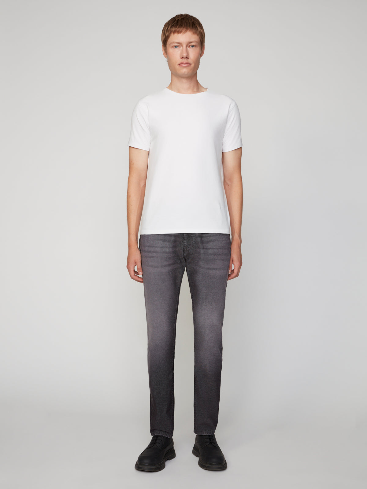 A person stands facing forward against a plain light background, wearing a white short-sleeve t-shirt, black shoes, and PROJEK RAW MENS BARU5-POCKET STRAIGHT FIT DENIM PANT in dark gray.