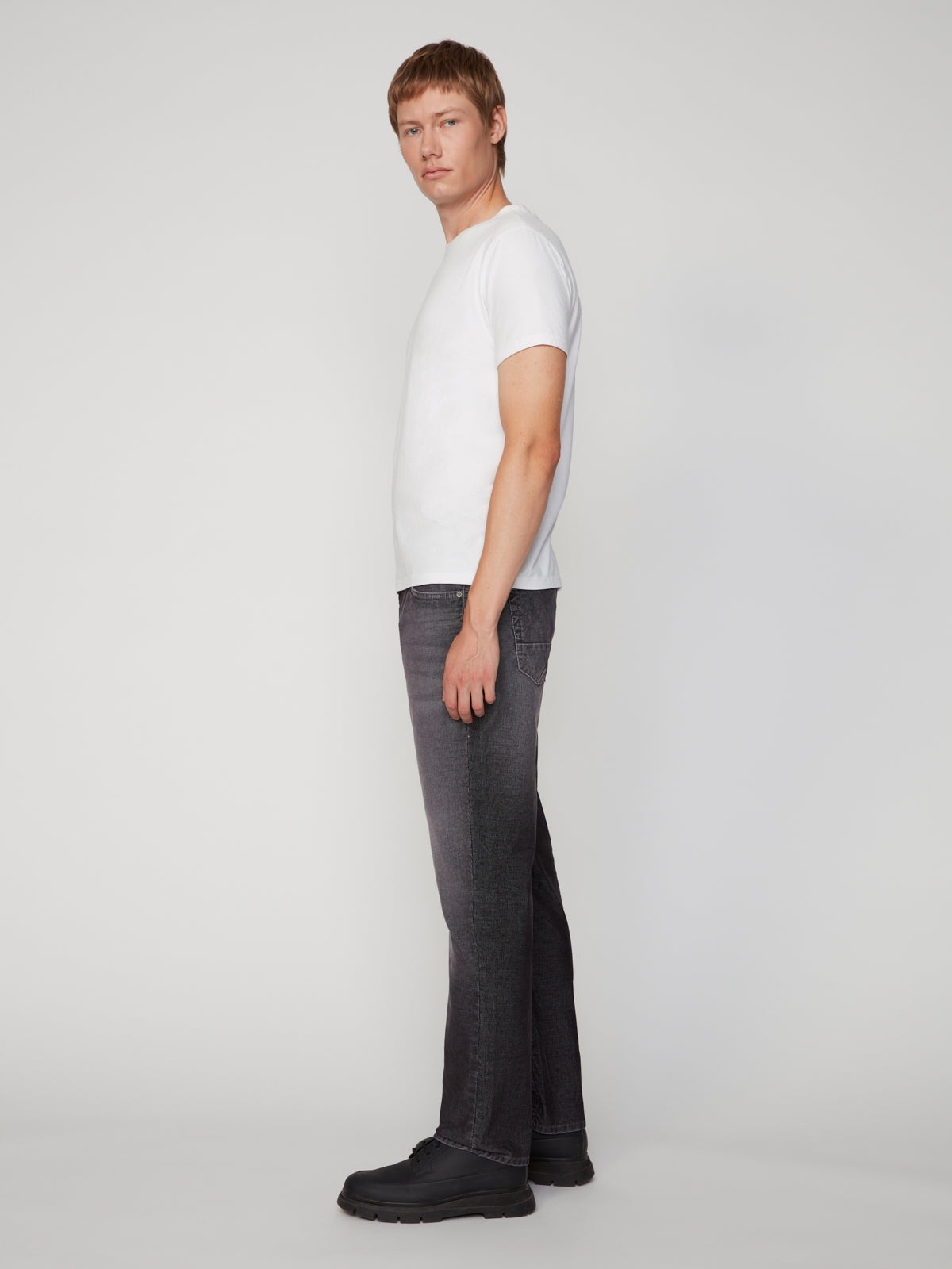 A young man stands in profile against a plain background, wearing a white t-shirt, black shoes, and PROJEK RAW’s MENS BARU5-POCKET STRAIGHT FIT DENIM PANT.