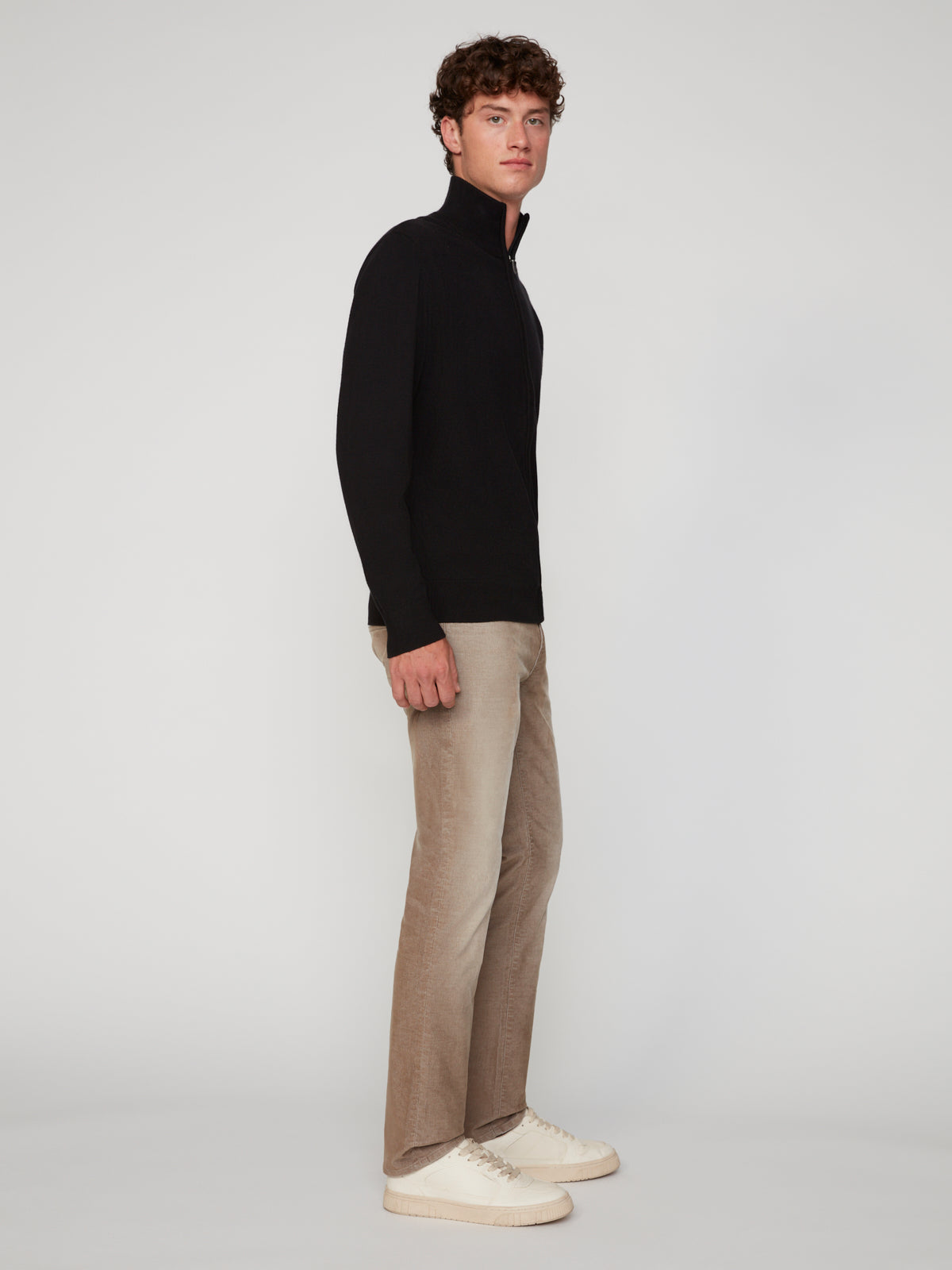 A young man stands sideways against a plain background, wearing a black zip-up sweater, PROJEK RAW MENS BARU5-POCKET STRAIGHT FIT DENIM PANT, and white sneakers. He looks slightly toward the camera with a neutral expression.