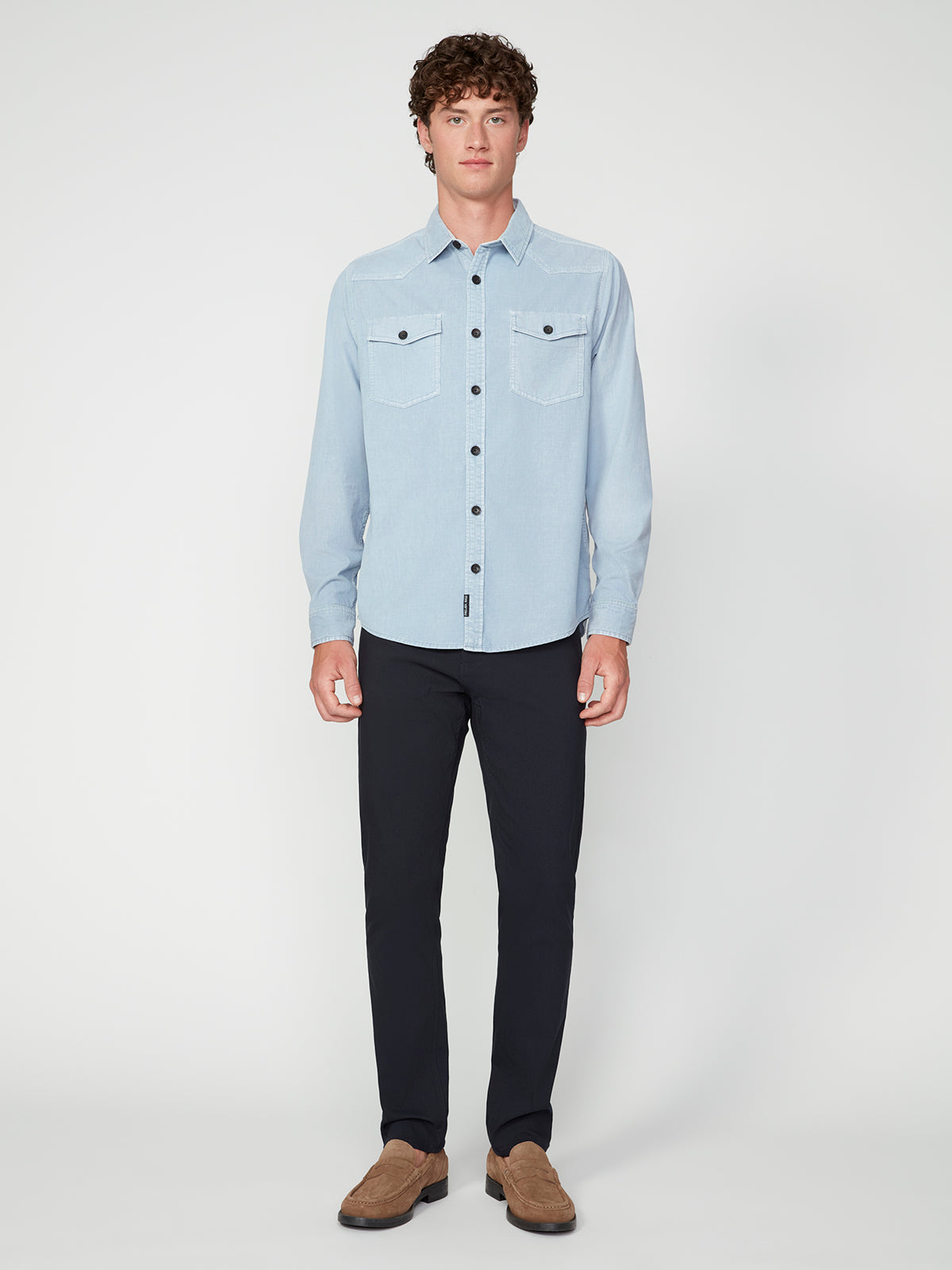 A young man with curly brown hair models the PROJEK RAW MENS LONG SLEEVES CORDUROY SHIRT in light blue, featuring chest pockets, paired with dark pants and brown shoes against a plain white background.
