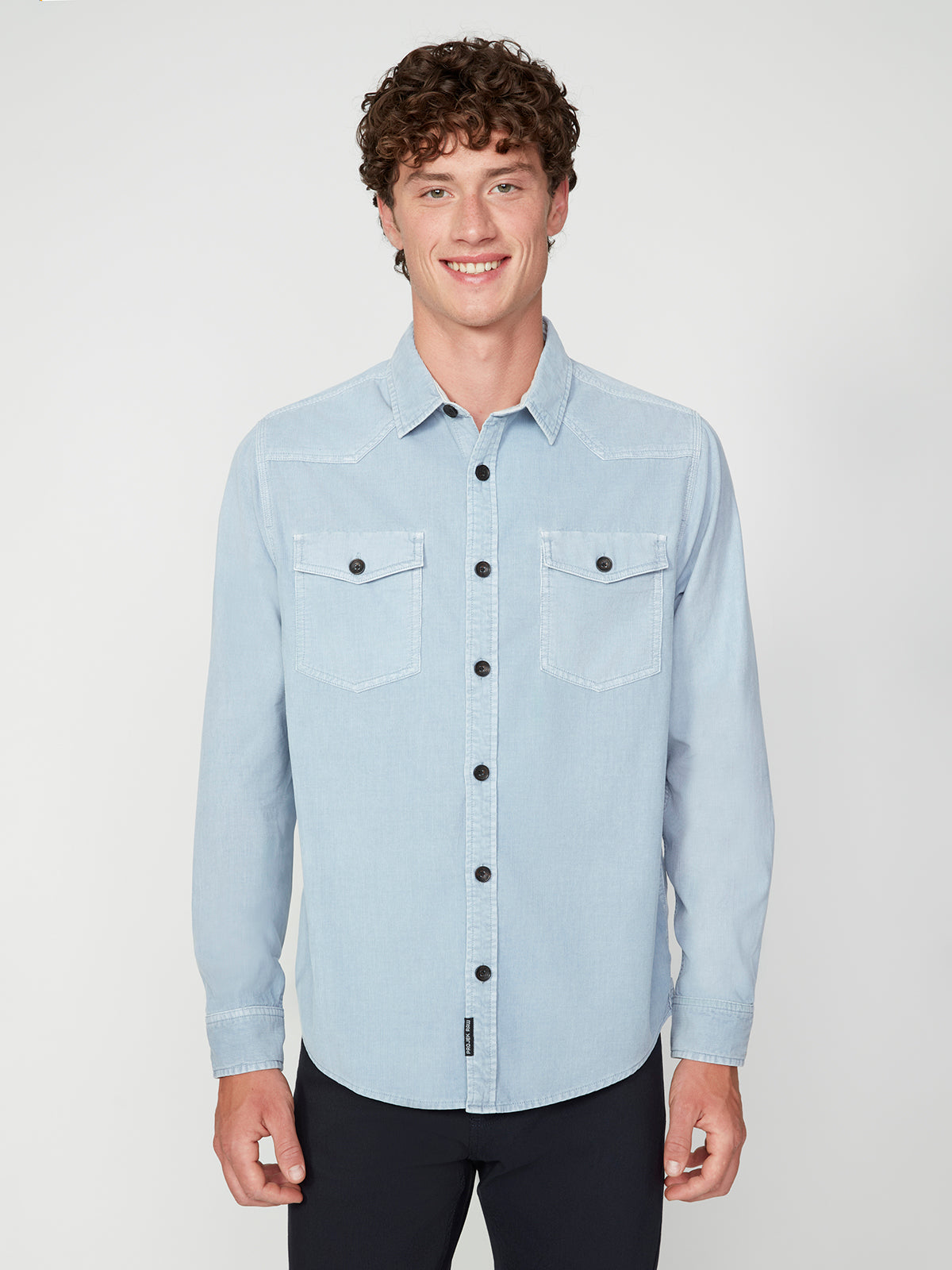 A young man with curly brown hair smiles at the camera, wearing the PROJEK RAW MENS LONG SLEEVES CORDUROY SHIRT in light blue with two chest pockets and black pants, set against a plain white background.