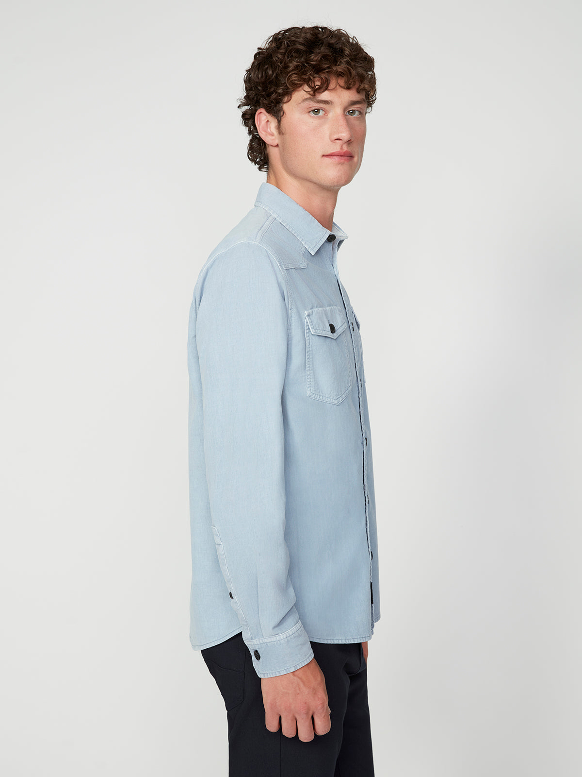 A young man with curly hair stands in profile against a plain background, wearing the PROJEK RAW MENS LONG SLEEVES CORDUROY SHIRT in light blue with front pockets, paired with dark pants.
