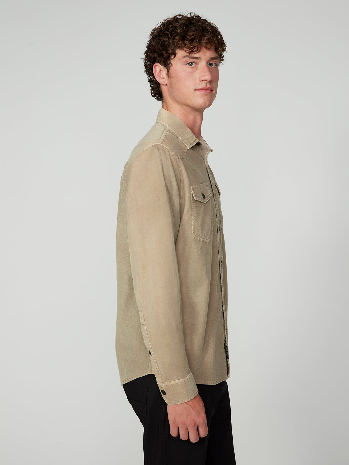 A young man with curly brown hair stands sideways facing left, wearing a PROJEK RAW MENS LONG SLEEVES CORDUROY SHIRT in light tan with chest pockets and black pants against a plain light gray background.