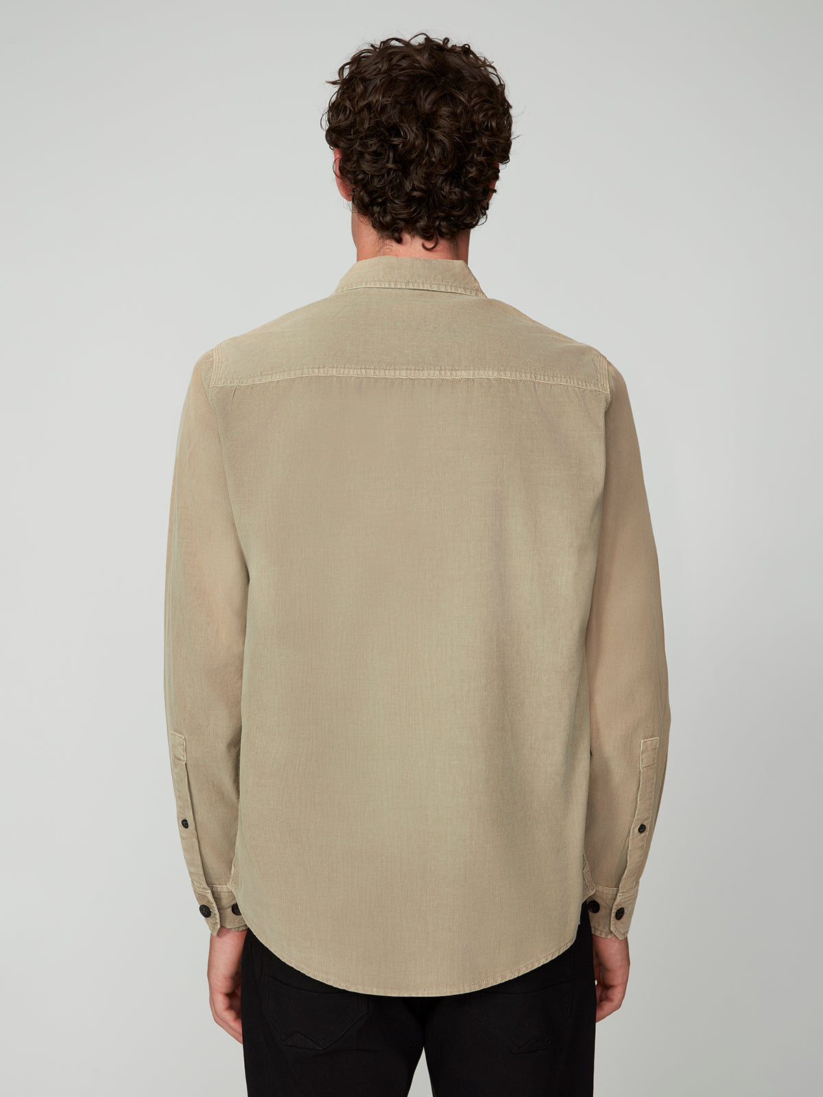 A person with curly brown hair is seen from behind, wearing the PROJEK RAW MENS LONG SLEEVES CORDUROY SHIRT in light beige with black buttons and black pants, set against a plain light background.