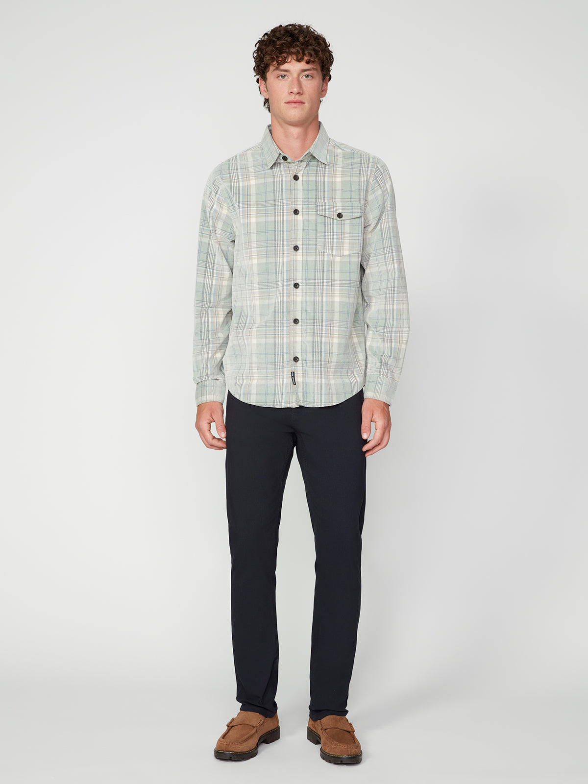 A young man with curly hair faces forward, wearing the PROJEK RAW MENS LONG SLEEVES PLAID CORDUROY SHIRT, paired with dark pants and brown shoes against a white background—perfect for enhancing any casual wardrobe.