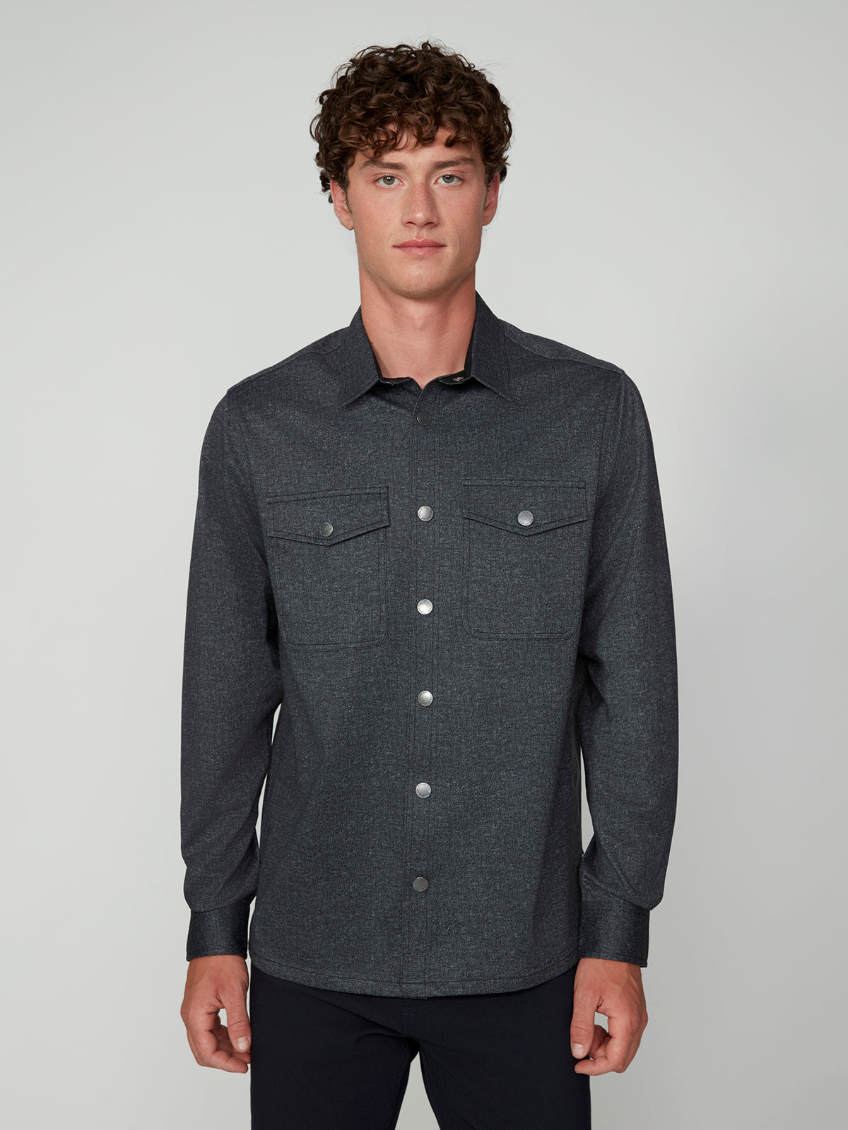 A young man with curly brown hair is wearing a dark gray PROJEK RAW MENS LONG SLEEVES KNIT SHIRT with two chest pockets, standing against a plain light gray background.