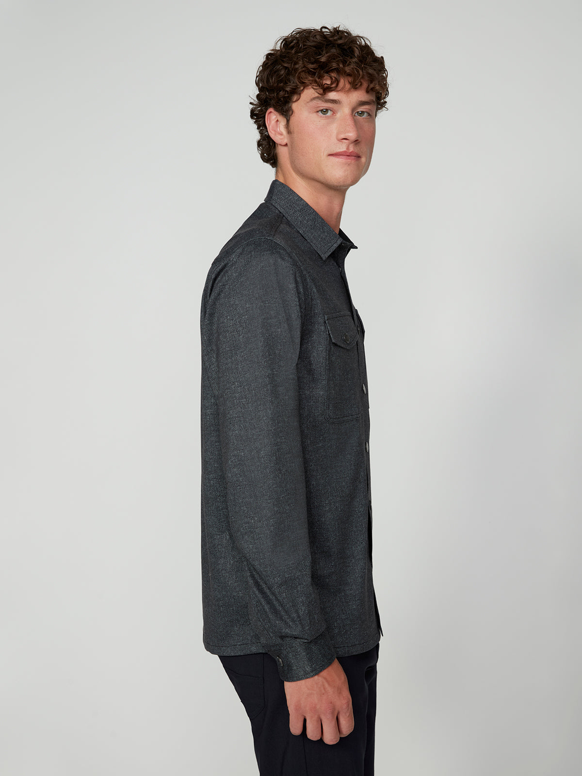 A young man with curly brown hair stands in profile, wearing a dark gray PROJEK RAW MENS LONG SLEEVES KNIT SHIRT and black pants, against a plain light gray background.